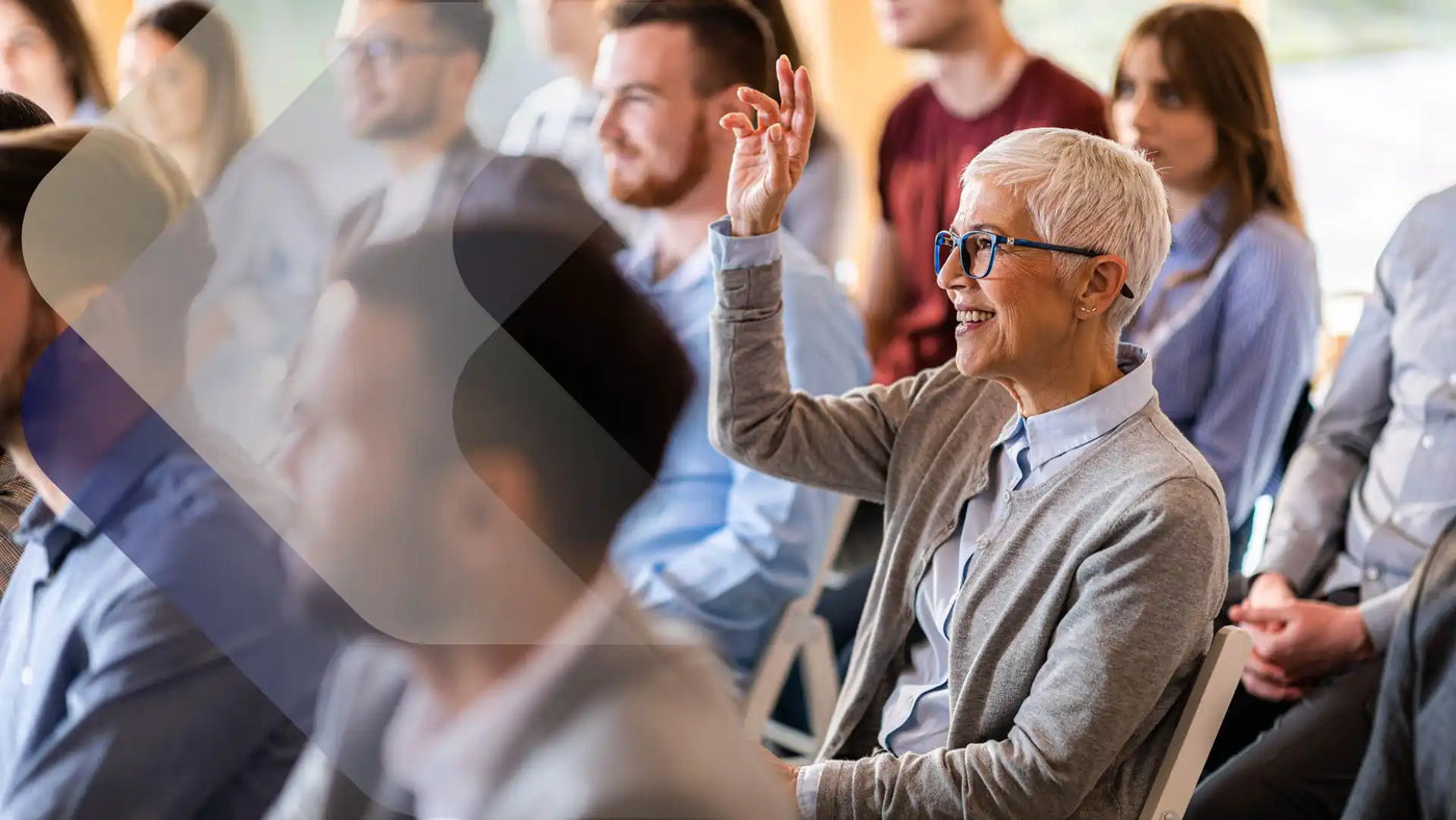 Lächelnde ältere Frau mit weißen Haaren und Brille, die in einer Gruppensituation die Hand hebt.